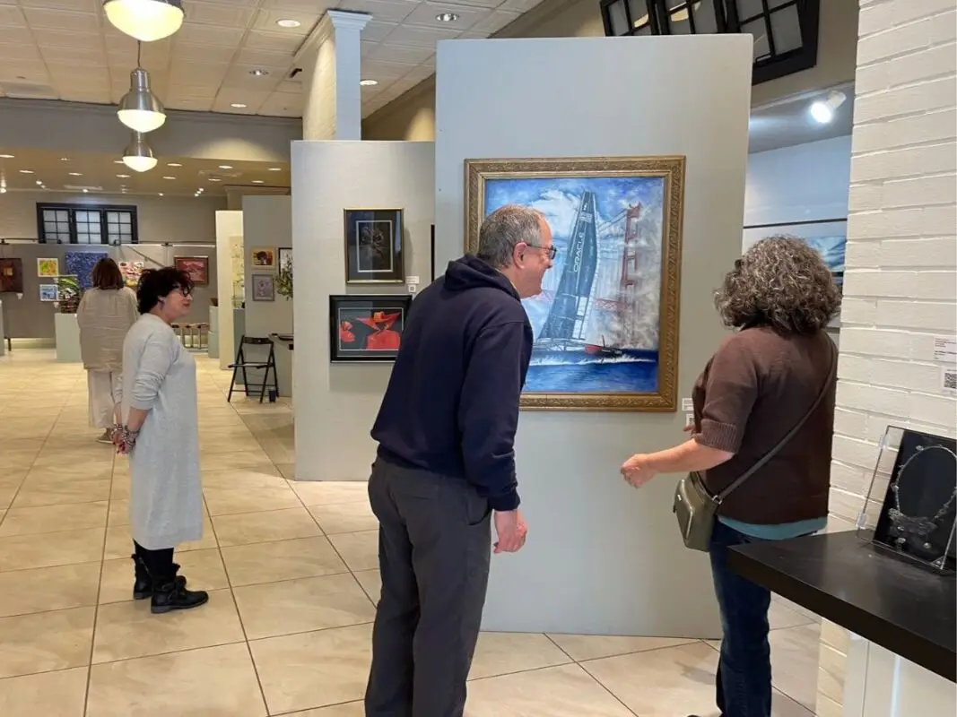 Two people in a gallery admire a framed painting on a light-colored wall, depicting a boat in the water. Other artworks hang around them. A woman wearing a white outfit observes other pieces in the background. Visiting this gallery is one of the best things to do in North Bay in May, with its spacious and bright interior.