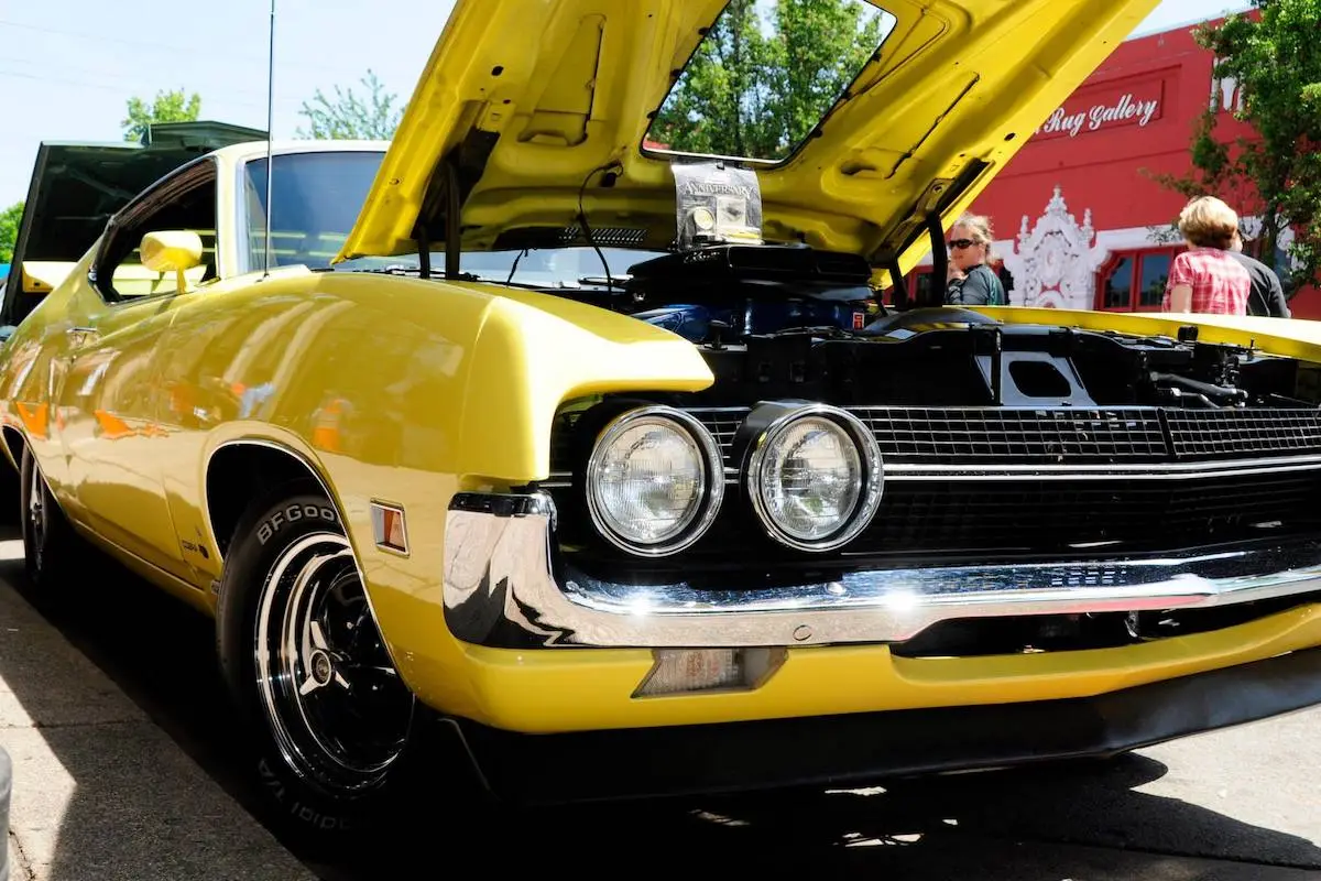 A classic yellow and black muscle car with its hood open is parked outdoors on a sunny day. People and trees are visible in the background, along with a red building displaying the name "Art Gallery." Checking out local car showcases like this is one of the best things to do in North Bay in May.