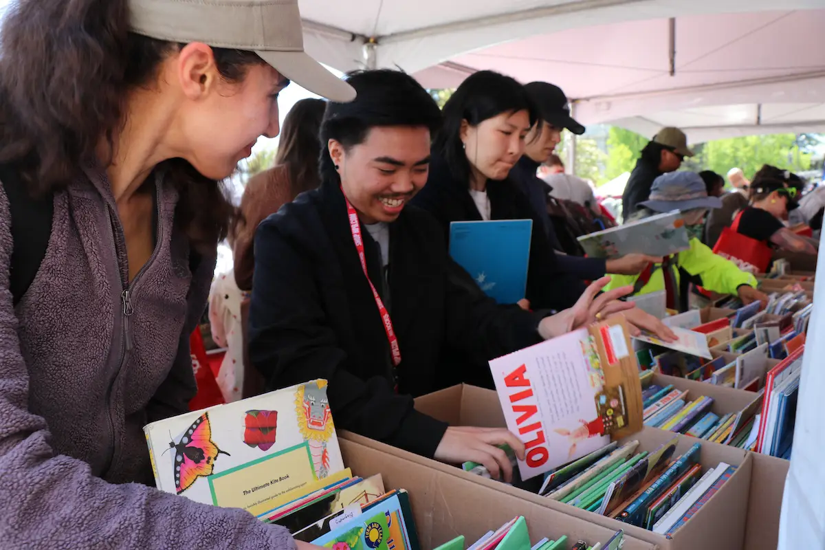 A group of smiling people browse through boxes of colorful children's books under a white tent at an outdoor event. One person holds a book titled "Olivia." The atmosphere is cheerful and busy as people search for books, making it one of the best things to do in the East Bay this June.