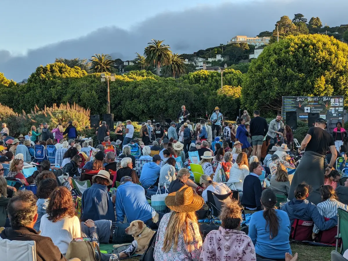 A large crowd is gathered outdoors on a grassy area, sitting on blankets and chairs for a live music event—one of the best things to do in North Bay in June. The stage is surrounded by trees and hills, as people enjoy the atmosphere and listen to the band. A dog lies contentedly among the attendees.