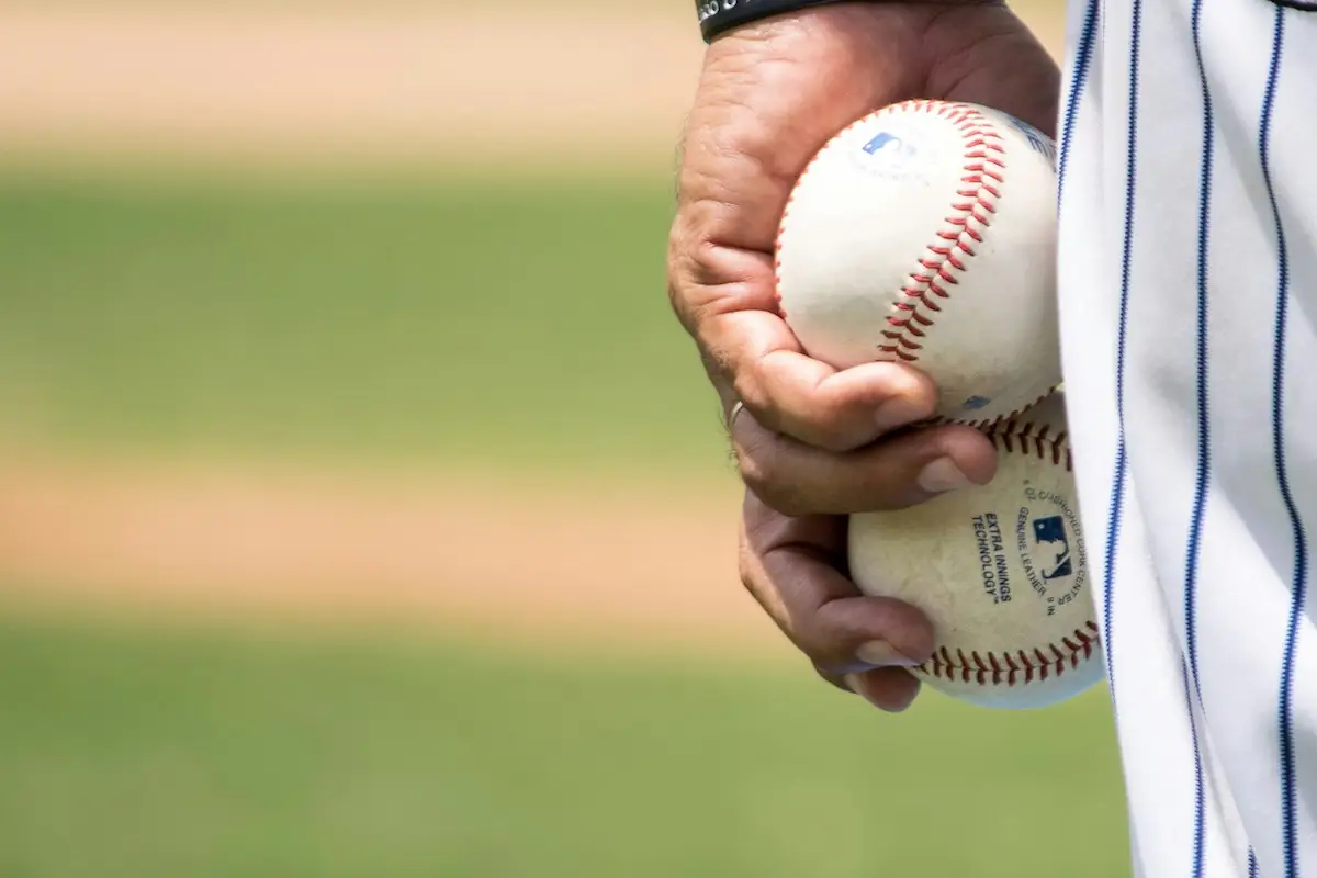 A person in a pinstriped uniform holds two baseballs in their left hand, preparing for a game. The background shows a blurred view of a grassy field, reminiscent of the popular sports events among the best things to do in East Bay this June.