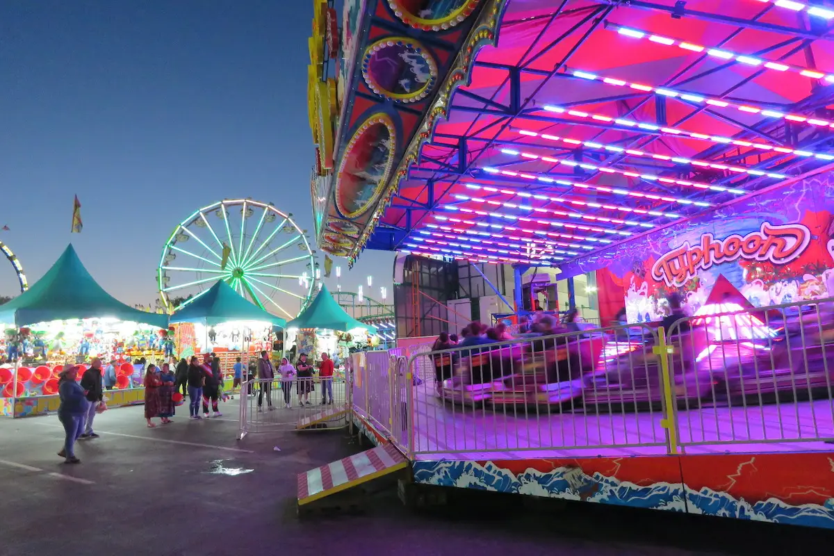 A vibrant carnival at dusk features a brightly lit ride named "Typhoon," with neon pink and blue lights. In the background, a large Ferris wheel and several green-topped tents are visible. One of the best things to do in South Bay this June, many people are enjoying the various attractions.