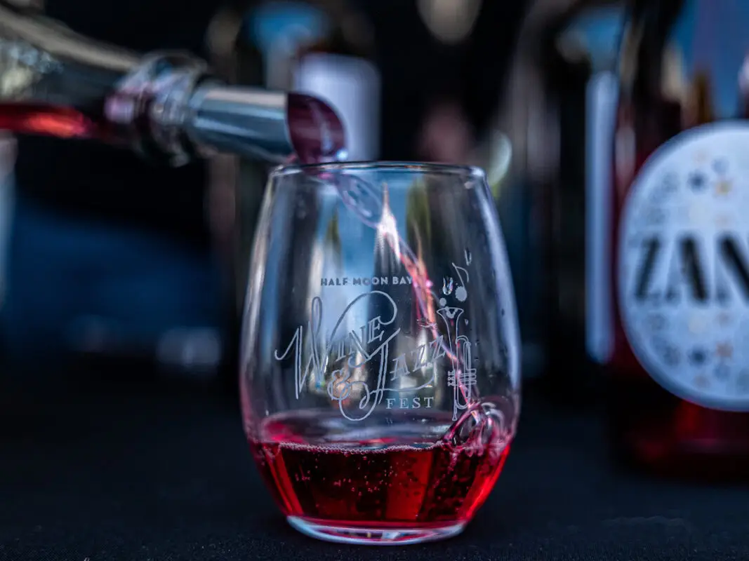 Close-up of a glass being filled with red wine at the Half Moon Bay Wine & Jazz Fest, one of the best things to do in South Bay this June. The event's logo adorns the glass, while blurred wine bottles in the background keep the focus on the pour and the glass.