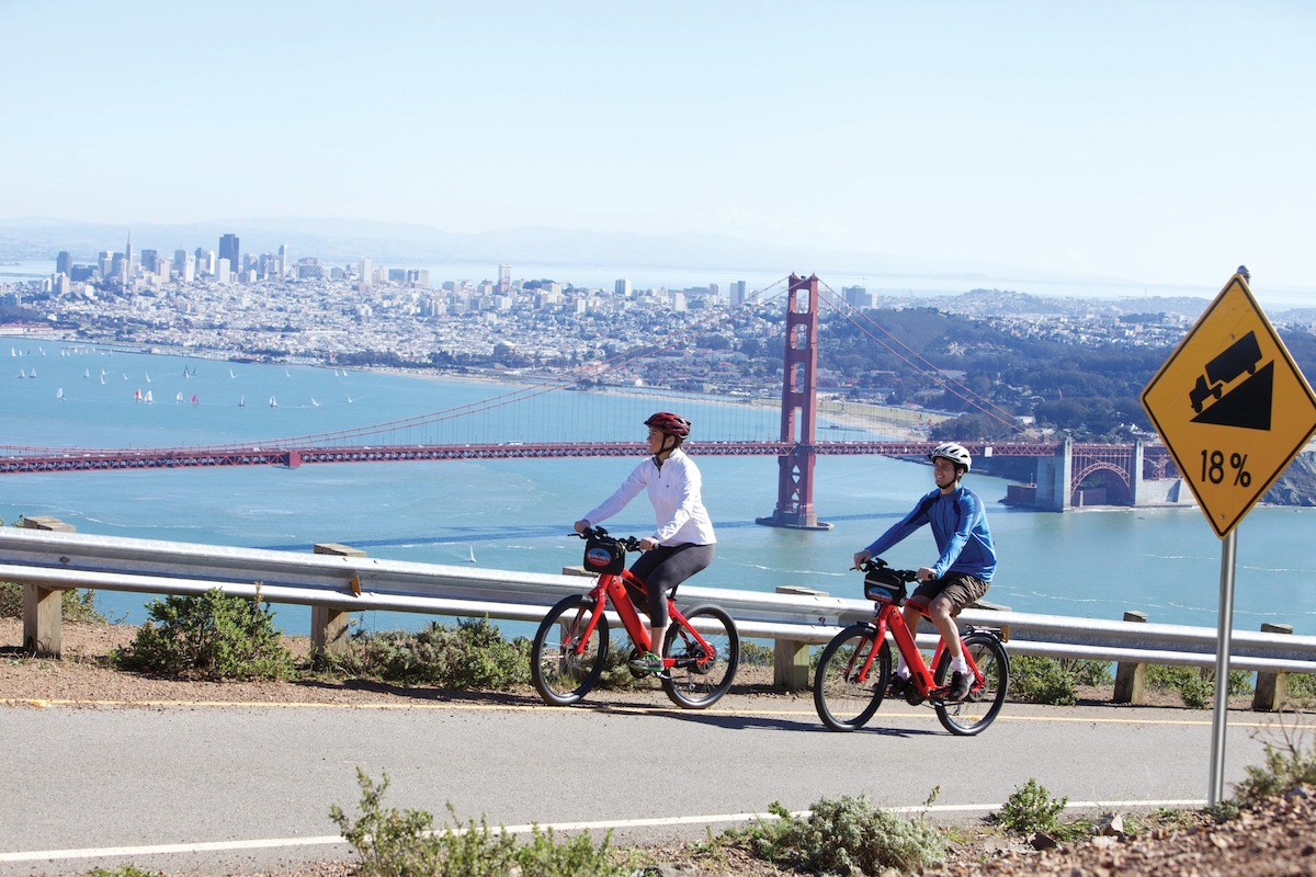 Two bikers bike uphill with a view of the bay, the Golden Gate bridge and San Francisco in the background.