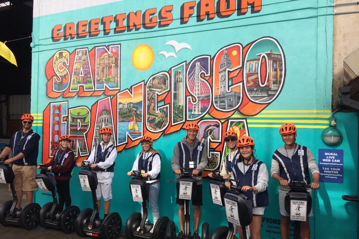 A group of tourists on segways wearing helmets stand in front of a mural that reads "Greetings from San Francisco."