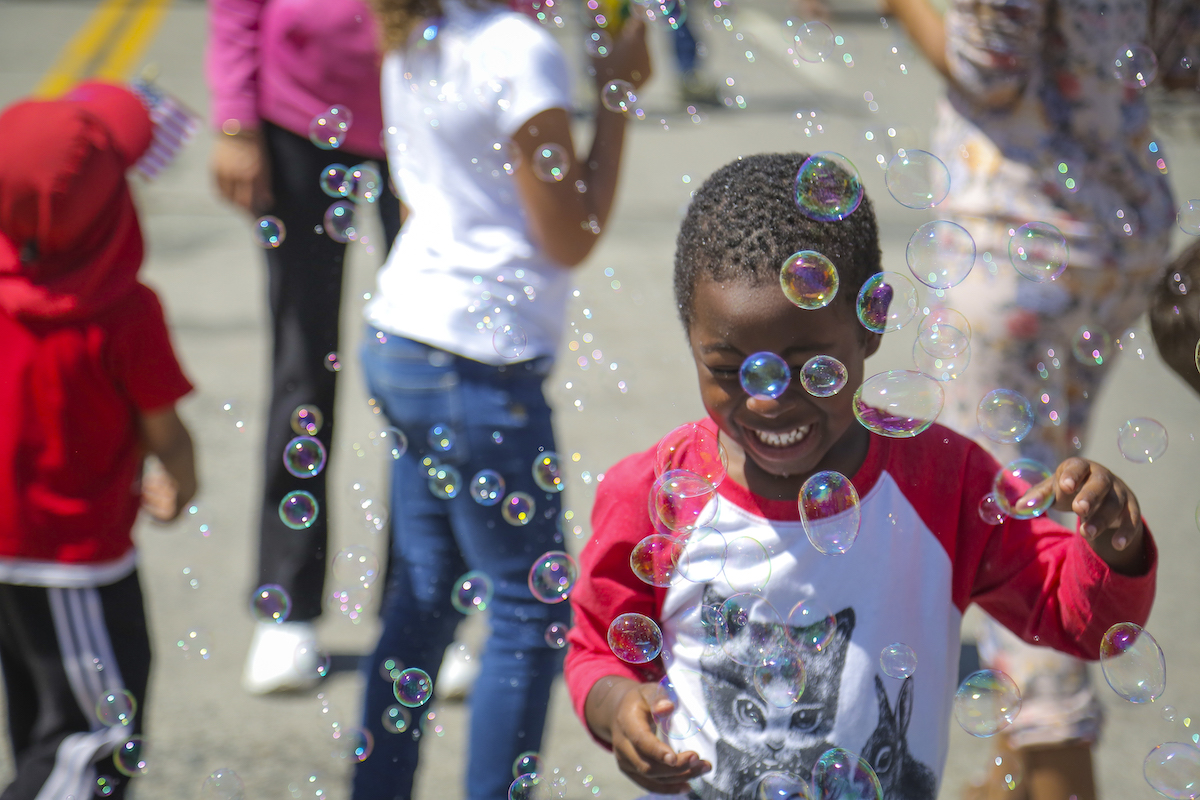 A young child joyfully playing with bubbles at an outdoor event, showcasing one of the best things to do in the Bay Area this August. The child is wearing a red and white shirt with a graphic on it. Several other people, including a person in a red hoodie, are also visible in the background enjoying the sunny day.