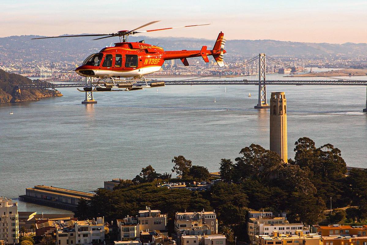 A red helicopter flies over San Francisco at dusk with a view of the Bay Bridge in the background.