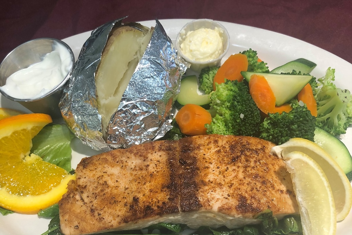 A plate with some cooked meat, cooked spinach, broccoli/carrots/cucumber, a baked potatoe, butter, sour cream and some citrus slices.