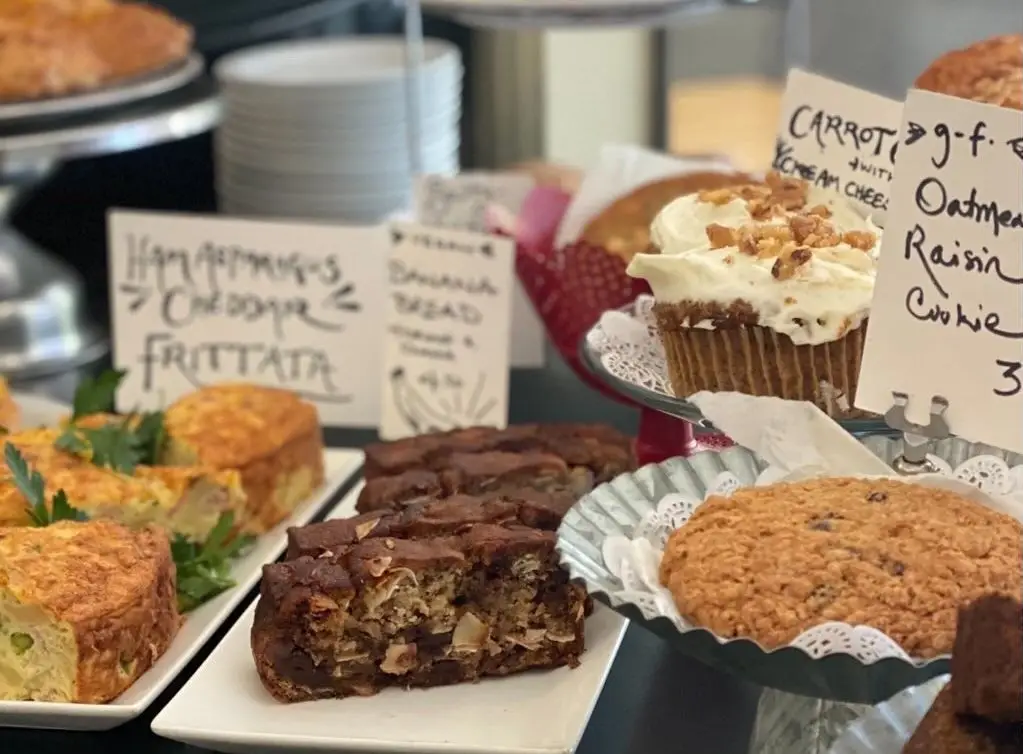 A variety of baked goods displayed on a table, including muffins, oatmeal raisin cookies, frittata slices, banana bread, and carrot cake. Handwritten signs indicate gluten-free options. Plates and a napkin holder are in the background—truly offering the best breakfast in Sonoma County.