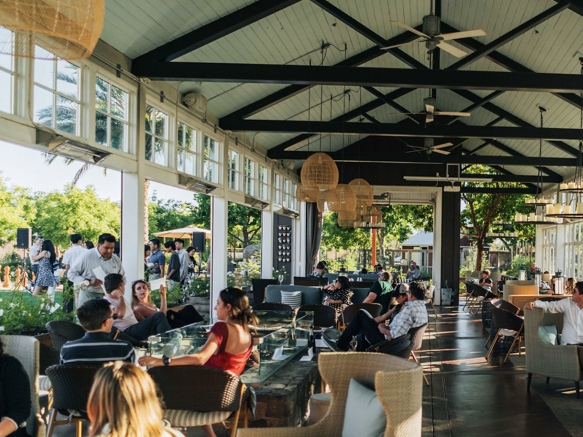 People are dining in a spacious, open-air restaurant with high ceilings and large windows. The room is filled with natural light, wicker furniture, and hanging lamps. Outside, lush greenery enhances the relaxed atmosphere—a perfect spot to explore among the Best Things to Do in Napa Valley This September.