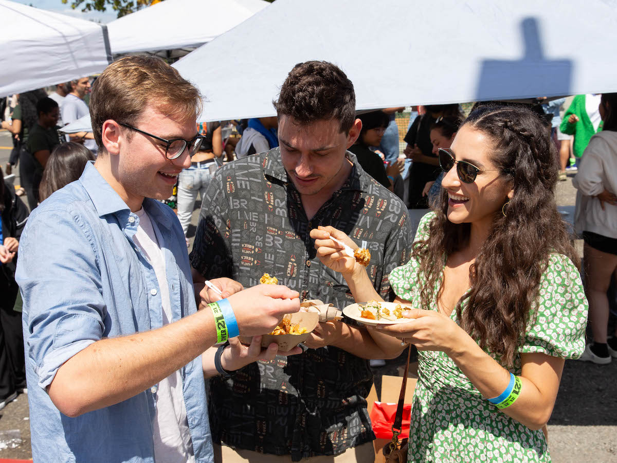 Three people standing in an outdoor market or festival, each holding a food plate. They appear to be enjoying their food and engaging in conversation. One person wears a blue shirt and glasses, another wears a patterned shirt, and the third person wears a green dress and sunglasses—definitely one of the best things to do in the East Bay this September.