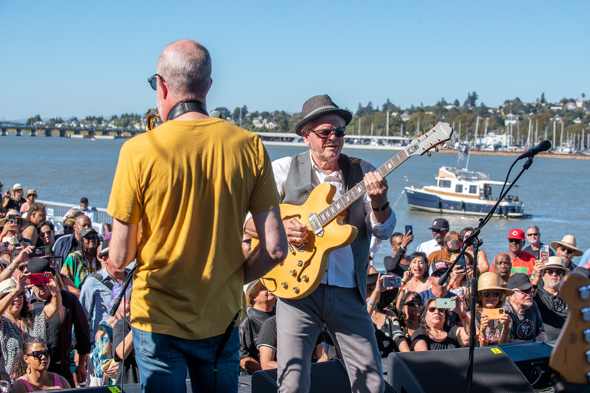 A guitarist wearing a hat and sunglasses plays on an outdoor stage with a lively crowd in the background. Another musician in a yellow shirt is visible from behind. The scene, one of the best things to do in the East Bay this September, is set by a waterfront with boats and a bridge under a clear blue sky.