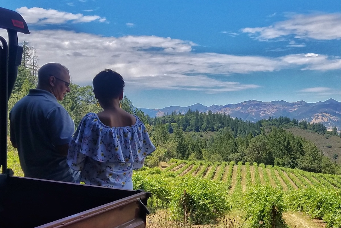 A man and a woman stand in front of a red truck overlooking a vineyard.