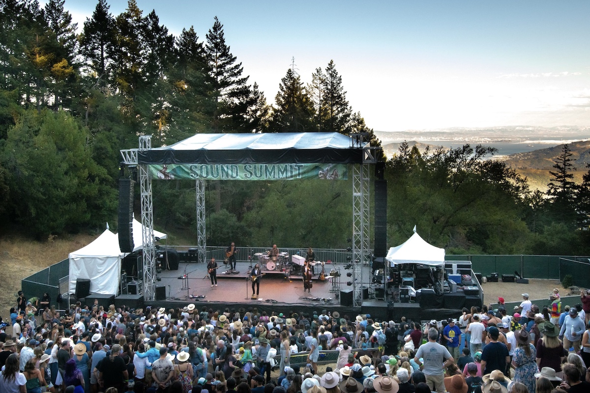 A crowd gathers in an outdoor amphitheater surrounded by trees, enjoying a band perform on stage with "Sound Summit" displayed on the canopy. Behind the stage, a scenic view of the landscape and water is visible under a clear sky—a perfect example of the Best Things to do in North Bay this September.