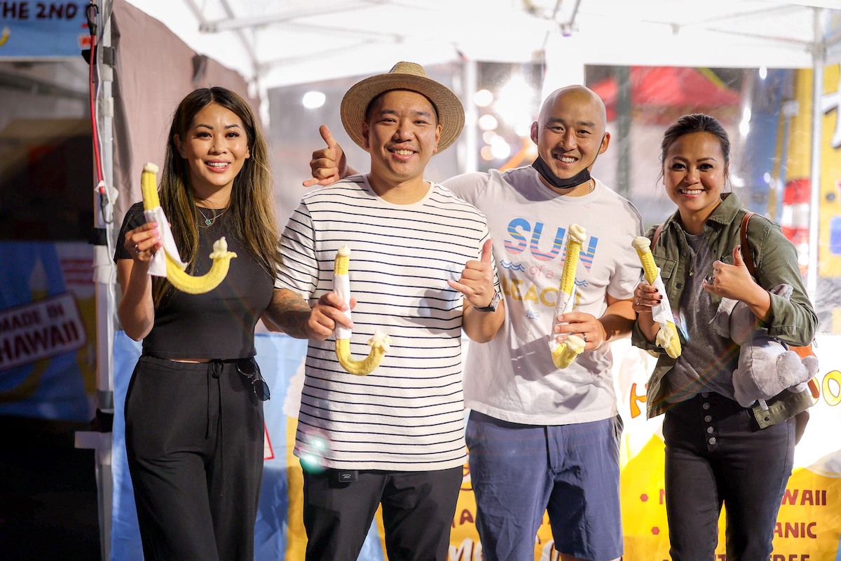 Four people standing together in a festive outdoor setting, smiling and holding large, curved food items. Two men in the middle wear casual shirts, while two women on the ends dress neatly. A stall tent and colorful decorations create a vibrant backdrop, showcasing some of the Best Things to do in the South Bay this August.