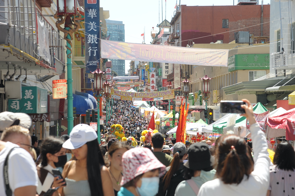 A bustling street market with colorful banners and tents lining the way is one of the best things to do in San Francisco this September. Crowds of people, some wearing masks and hats, walk through the market. Vibrant signage in both English and Chinese adorns the storefronts, creating a lively, multicultural atmosphere.