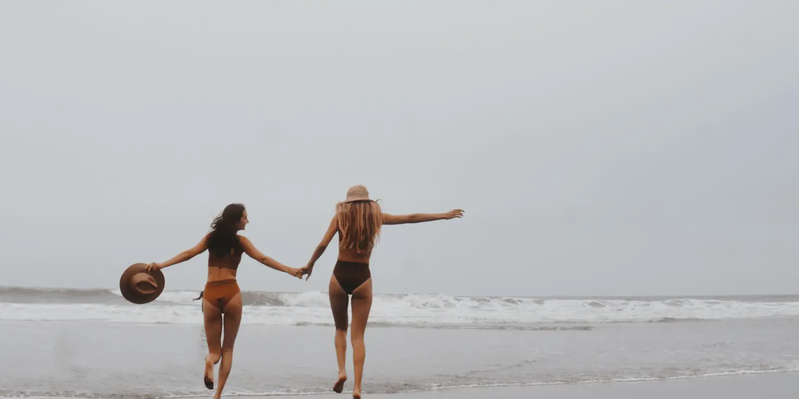 Two women in bikinis, holding hands, run towards the ocean on a cloudy day. One holds a hat, the other wears one. Waves crash gently in the background, creating a carefree and joyful beach scene perfect for a Shop Surf + Sand moment.
