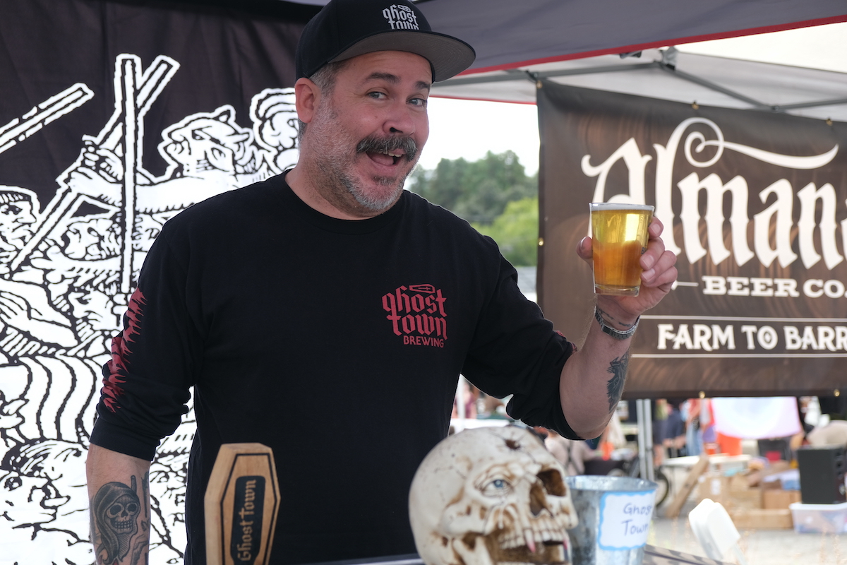 A person with a mustache and beard, wearing a black "Ghost Town Brewing" shirt and hat, smiles and holds up a glass of beer. They stand behind a booth decorated with a skull and "Almanac Beer Co." banner in the background—one of the best things to do in North Bay this October.