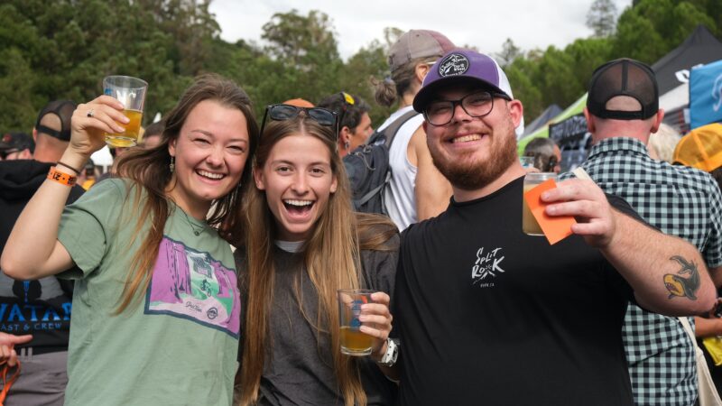 Three people smiling and holding drinks at an outdoor event, showcasing some of the best things to do in North Bay this October. The person on the left is wearing a green shirt, the person in the middle has long hair and sunglasses, and the person on the right sports a cap, glasses, and a black shirt. Trees and a lively crowd are visible in the background.