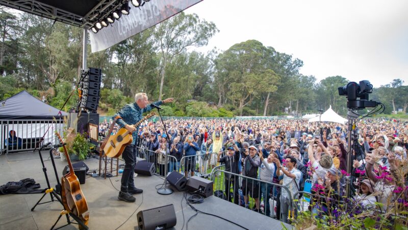 A musician on stage with a guitar points towards a large, energetic crowd during an outdoor concert—one of the Best Things To Do in San Francisco. People in the audience are cheering and raising their hands. Trees and tents are visible in the background under a clear sky.