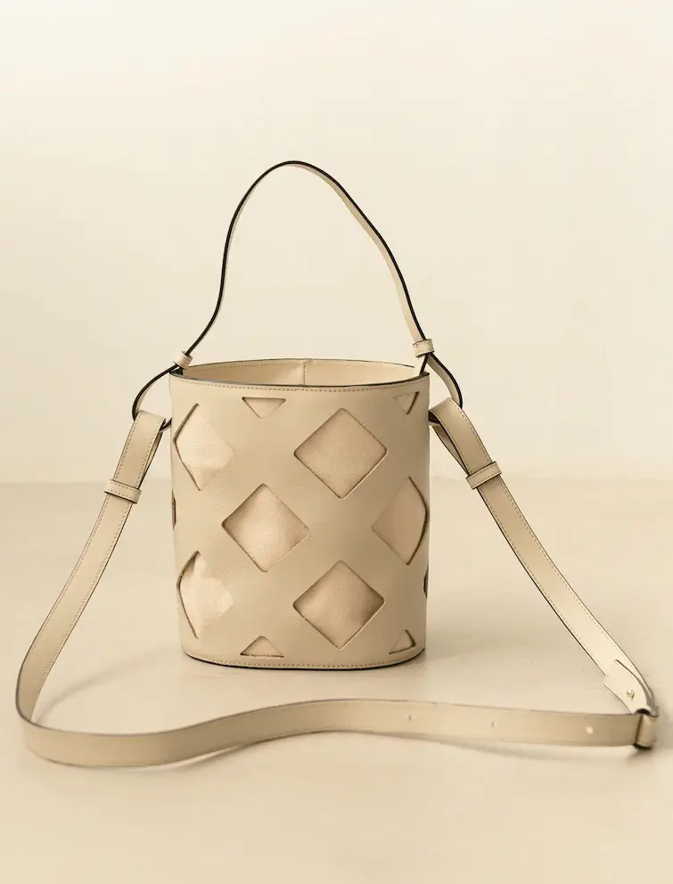 Discover the beige bucket-style handbag at Shop Surf + Sand, featuring a single top handle and an adjustable shoulder strap. This chic accessory boasts a distinctive geometric cutout pattern with diamond shapes, revealing a lighter inner layer. The plain, neutral background completes its elegant design.