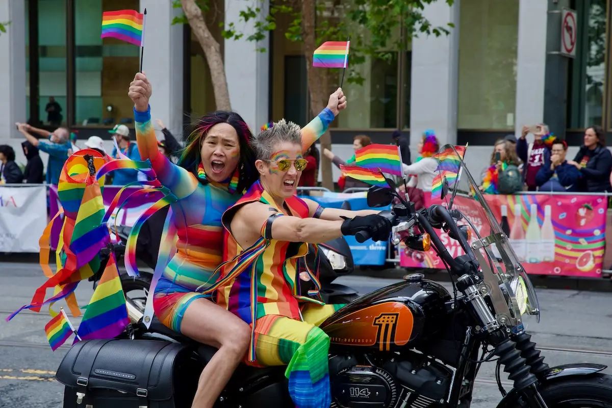 Two individuals wearing colorful rainbow-themed outfits ride a motorcycle while waving pride flags. They are surrounded by a spirited crowd celebrating at a Pride parade in San Francisco—one of the best things to do in June. The street is decorated with rainbow banners, and the atmosphere is festive and joyous.