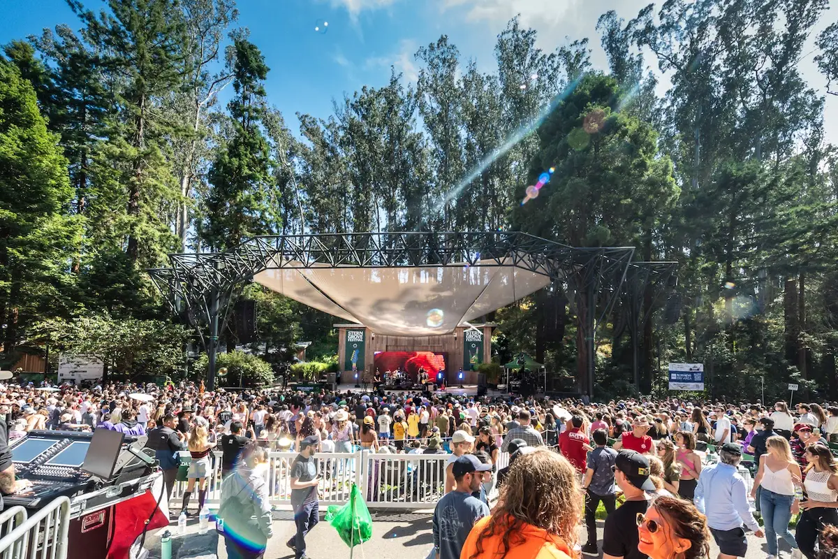 A large crowd enjoys an outdoor music festival in a forested area near San Francisco, one of the best things to do in June. Attendees stand in front of a stage sheltered by a tent-like structure, with performers visible on stage. Tall trees surround the festival scene, and the sky is clear and sunny.