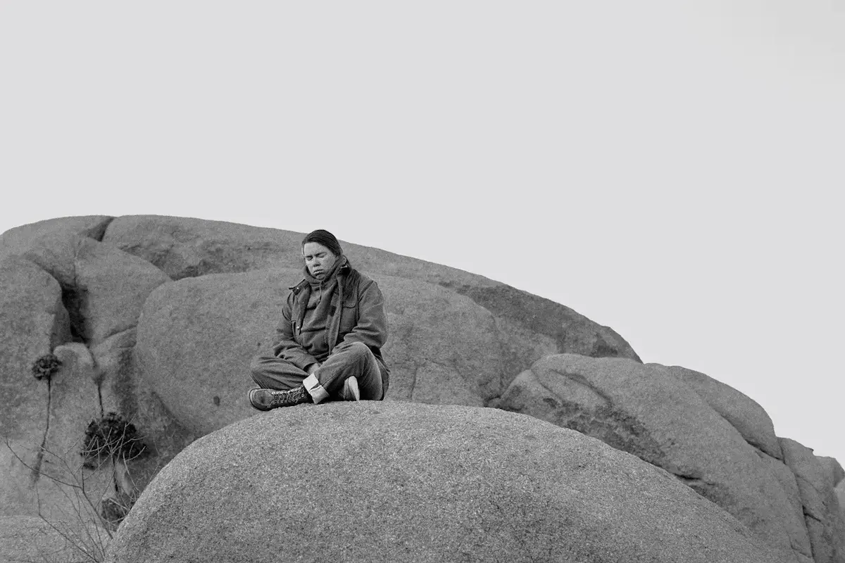 A person sits cross-legged on a large rock formation in San Francisco, wearing a jacket and pants, with a neutral expression. The backdrop of more large rocks against a clear sky enhances the serene and contemplative feel—truly one of the best things to do in June.