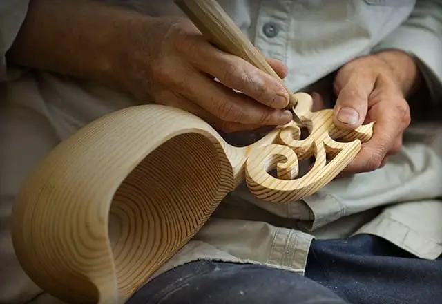 A person wearing a light shirt is intricately carving a wooden object with a handheld carving tool, showcasing some of the best things to do in Tahoe in November for craft enthusiasts. The carving features swirling patterns, and the wood grain is prominently visible, highlighting the detailed craftsmanship.