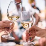 Two hands clink wine glasses filled with white wine in a celebratory gesture. In the blurred background, several people are seated and enjoying the occasion. The atmosphere appears relaxed and casual, possibly outdoors on a sunny day—one of the best things to do in the North Bay January.