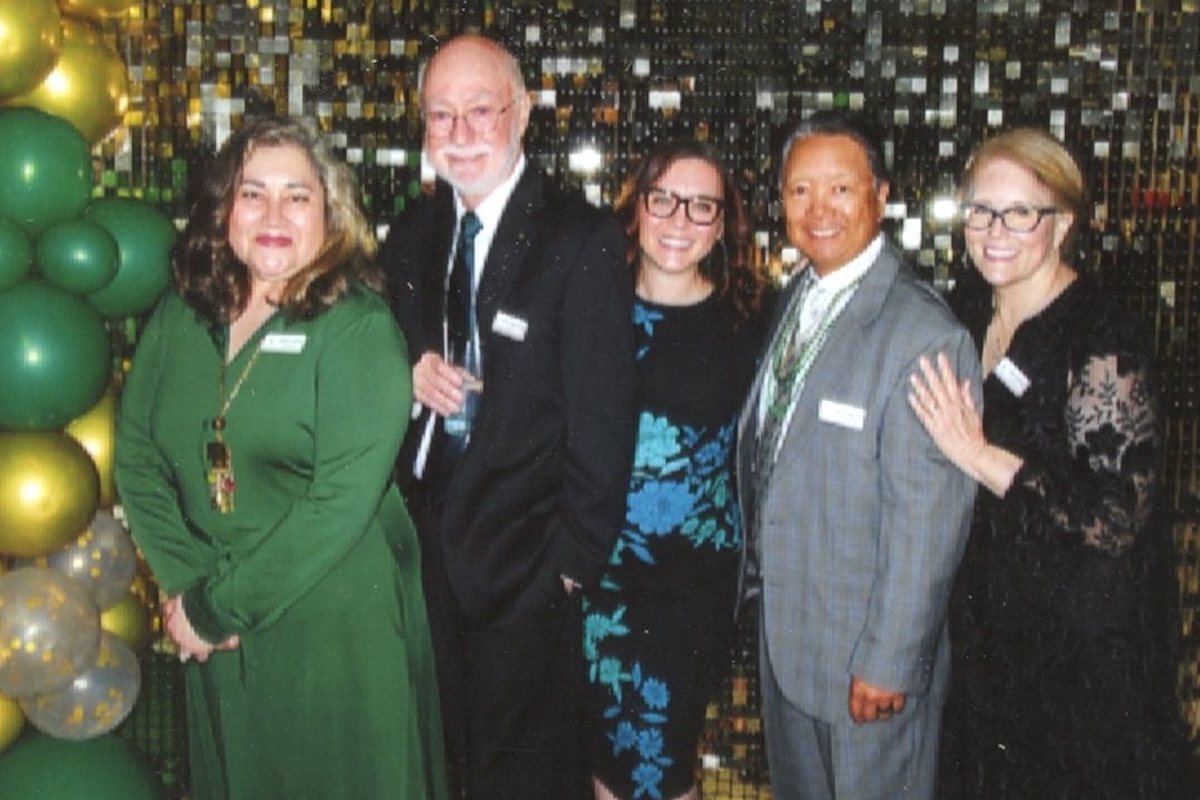 Five people in cocktail attire stand in front of a sparkly backdrop and next to a green and gold balloon pillar.