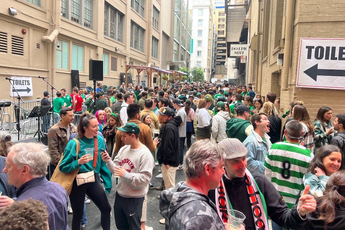 A large crowd of people standing in the streets of San Francisco wearing festive St. Patrick's day attire and holding drinks.