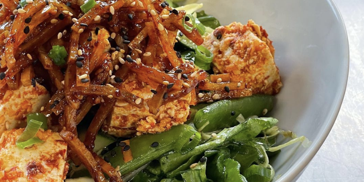 A bowl of vibrant food featuring a bed of greens topped with glazed tofu pieces, stir-fried vegetables, and a garnish of black and white sesame seeds, with a glistening sauce coating the dish—perfectly capturing the essence of enjoying the best dinner in Hilo on the Big Island.