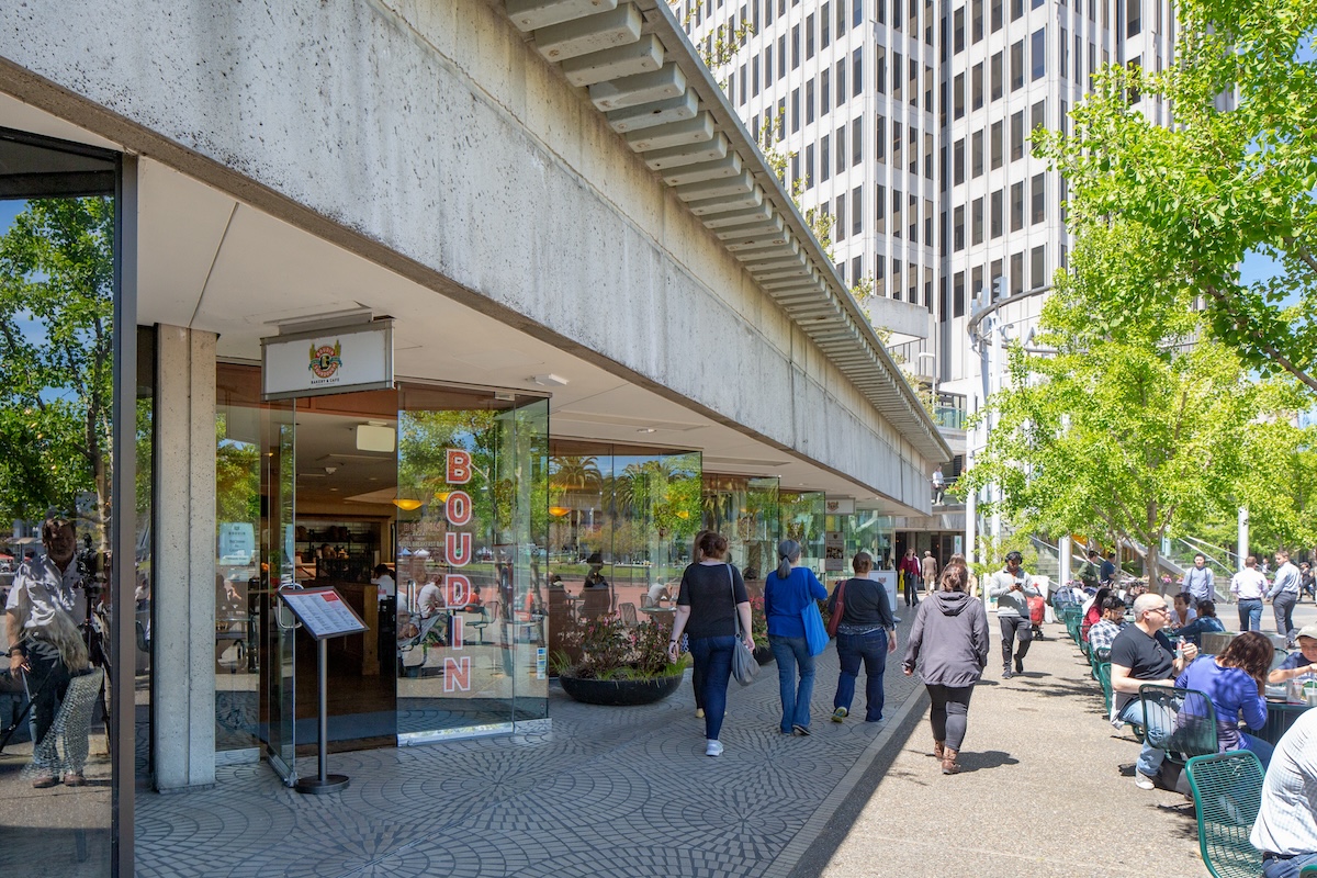 People walking along a sunny outdoor area near a Boudin bakery and cafe at the Embarcadero Center in San Francisco. The walkway, lined with trees and outdoor seating, leads to a tall building in the background. Patrons inside enjoy drinks by the glass entrance, where a menu stand is placed by the door.