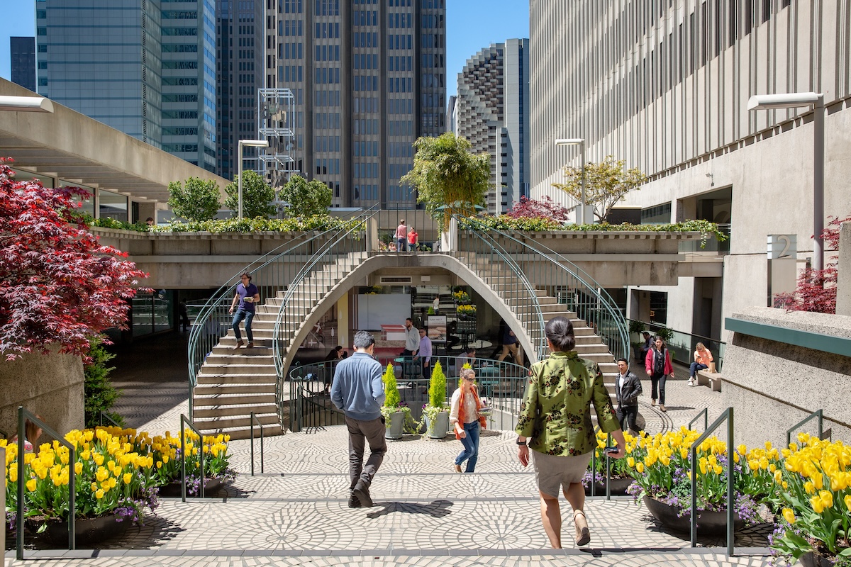 People walk on a patterned terrace featuring vibrant yellow flowers. A split staircase leads to an elevated walkway with potted plants and trees, reminiscent of the Embarcadero Center in San Francisco. Tall, modern buildings enclose the urban scene under a clear, blue sky.