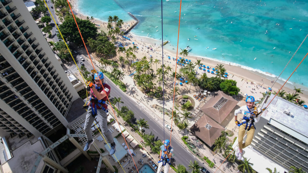 Three individuals participate in a rappelling activity down the side of a tall building, overlooking a scenic beach with turquoise waters, palm trees, and sunbathers. This adventure is one of the best activities to explore when considering what to do in Hawaii in October. The participants are equipped with safety gear and appear to be enjoying the thrilling descent.