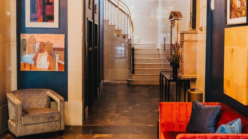 A modern hotel lobby in one of the top south bay hotels featuring dark blue walls adorned with colorful paintings. A red sofa and a gray armchair are positioned near the entrance. The floor is tiled, and in the background, a curved staircase with a wrought-iron railing leads to the upper floor.