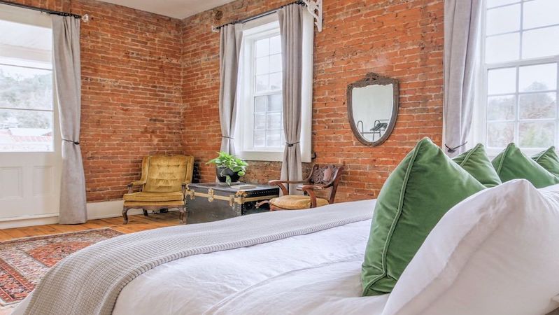 A cozy bedroom with brick walls features a bed with white linens and green accent pillows. The room, reminiscent of some charming South Bay hotels, includes large windows with light gray curtains, a vintage armchair, a trunk used as a coffee table, and a hanging mirror. A potted plant adds a touch of greenery.