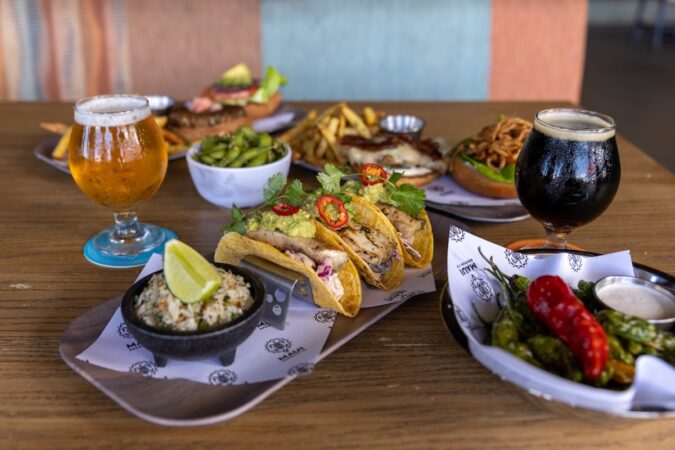 A table with tacos, sauce, edamame, peppers, other plates of food and a couple of glasses of cold beer.