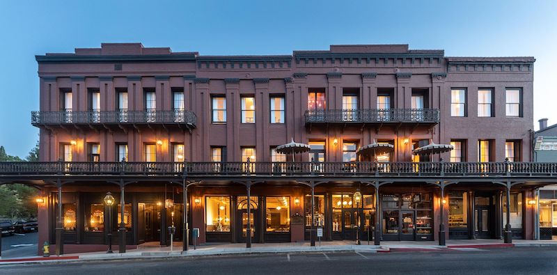 A broad, multi-story brick building illuminated with warm lights at dusk, perfect for those seeking where to stay in Sacramento. It features balcony railings on the upper floors and a covered sidewalk with railings on the ground level. The architecture is classic and well-maintained, evoking a historic feel.