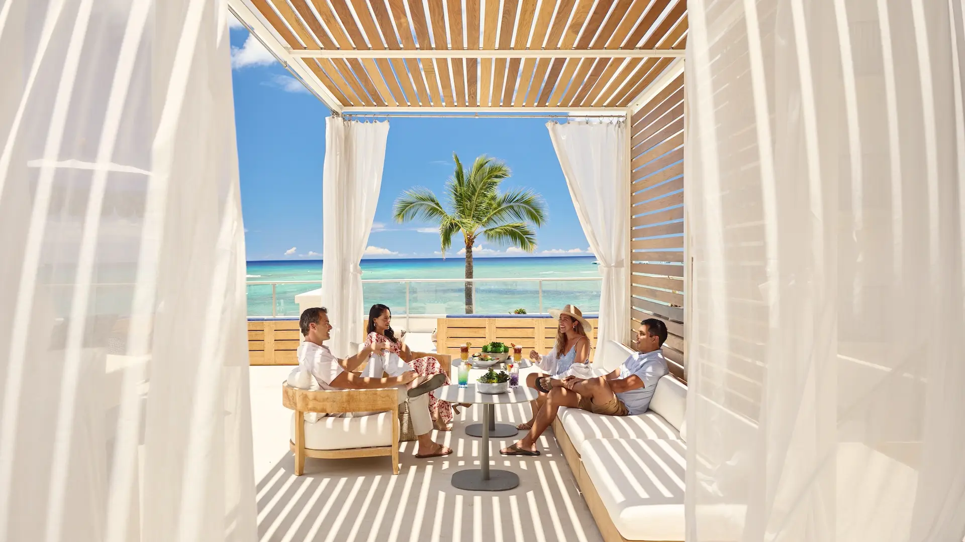 Four people relax in a shaded, open-air beach cabana with white curtains. They are seated on cozy furniture, enjoying drinks and conversation. In the background, there's a view of a palm tree, clear blue ocean, and sky. The setting exudes a serene, tropical feel—perfect for a local getaways sweepstakes.