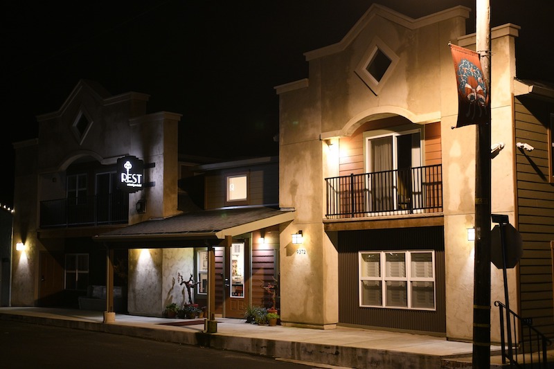 A street at night with two illuminated buildings; one features a sign that reads "REST," guiding travelers looking for where to stay in Sacramento. The building on the left has a covered entrance with lights, while the building on the right has a second-floor balcony and windows glowing warmly. A street light and banner are visible.