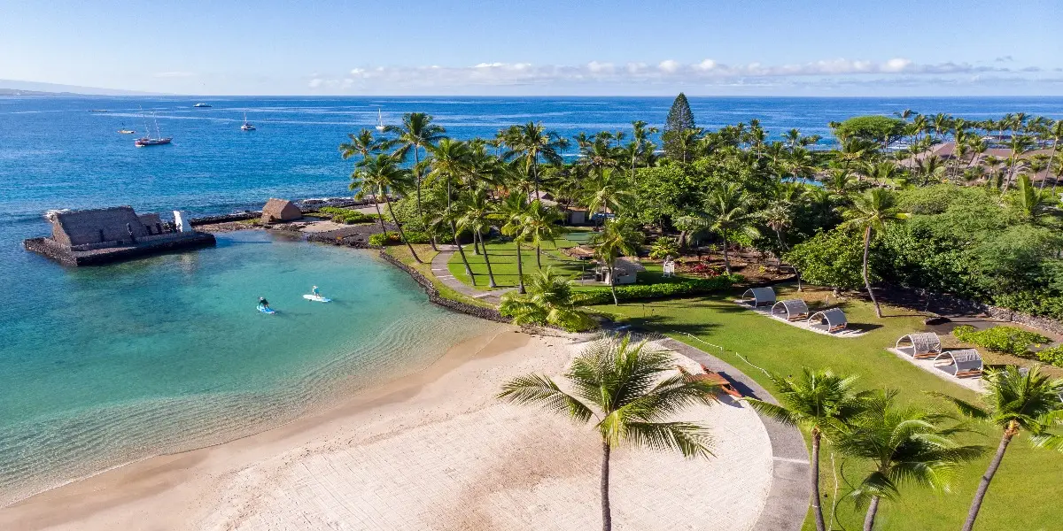 Aerial view of a tropical beach on the Big Island with clear blue waters and a white sandy shore. Palm trees dot the lush green landscape, with beach chairs and a small pavilion near the water. Kayakers paddle near the shore, boats anchored in the distance, offering views from some of the best vetted value hotels.