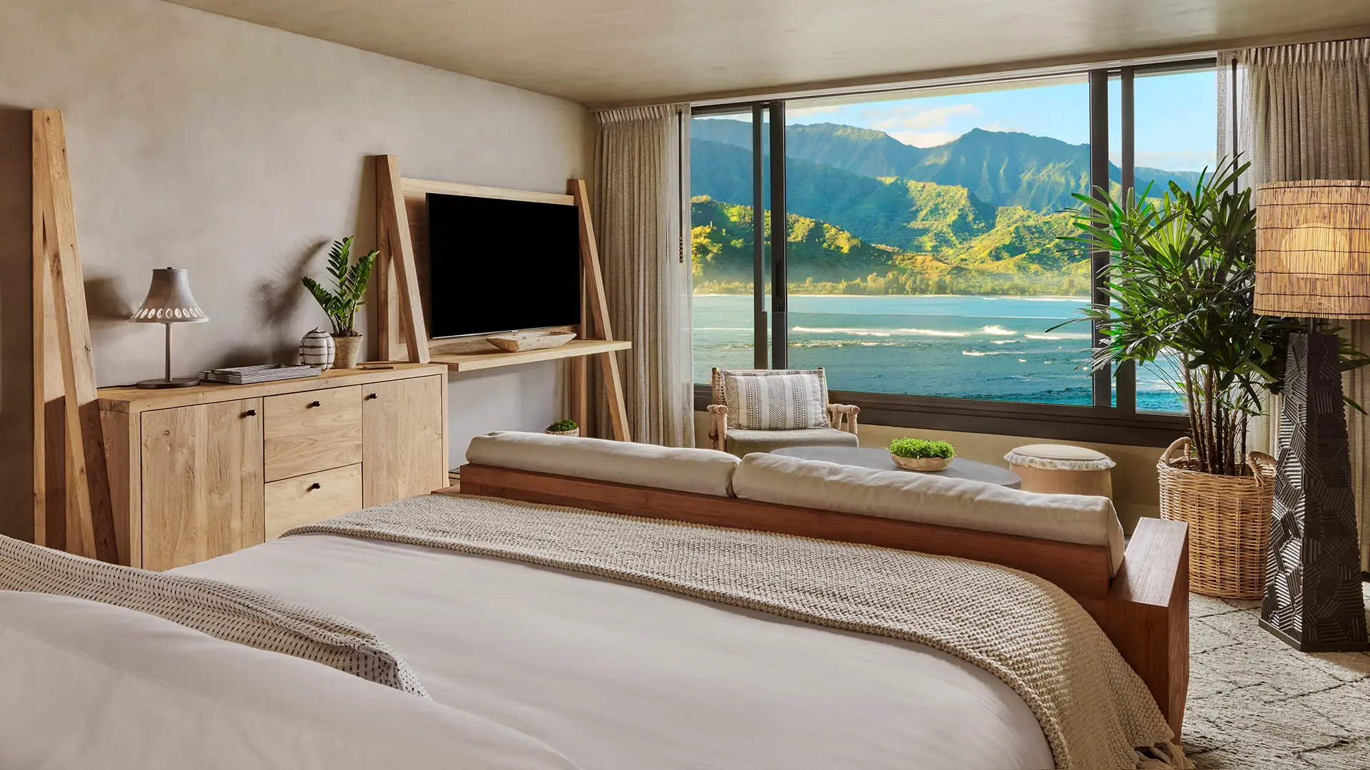 A well-lit bedroom with a large bed awaits you at one of the best romantic hotels in Kauai. The room features wooden furniture, a television, and a small plant. A large window offers a scenic view of mountains and water, as natural light floods the space, enhancing the cozy and serene atmosphere.