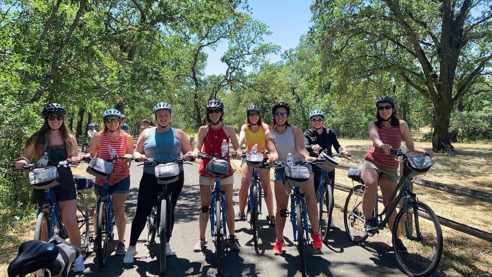 A group of eight people, all wearing helmets and casual summer clothes, are lined up on bicycles on a paved trail surrounded by trees and greenery. The sunny weather and shadows on the ground suggest it is a bright day, showcasing one of the top activities in Sonoma.