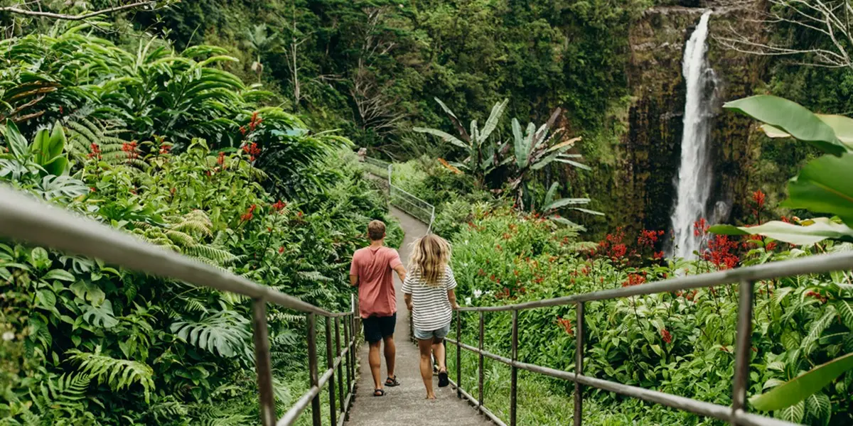 A couple walks down a pathway surrounded by lush greenery and vibrant red flowers toward a waterfall in a tropical setting, reminiscent of the best hikes on the Big Island. The man is wearing a pink shirt, and the woman is in a striped dress, embodying adventure and tranquility.