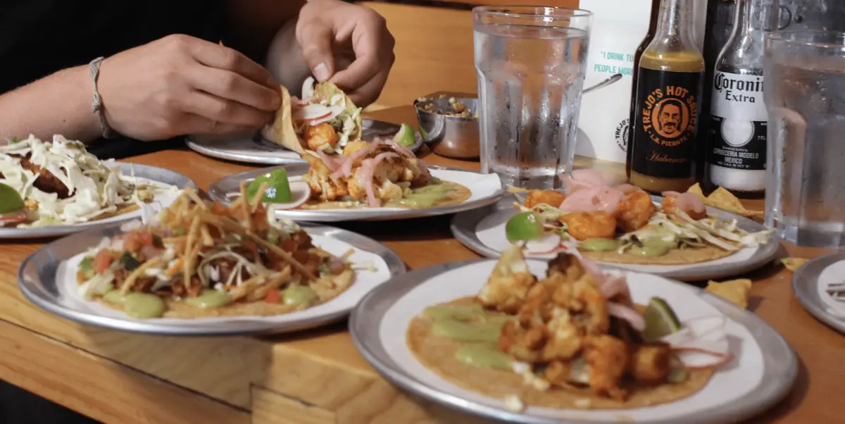 A table with multiple plates of tacos topped with various ingredients such as sliced radishes, cabbage, and sauce. Two hands are seen assembling a taco. There are beverages, including a bottle of hot sauce and a Corona Extra beer, and a glass of water on the table—a true taco feast.