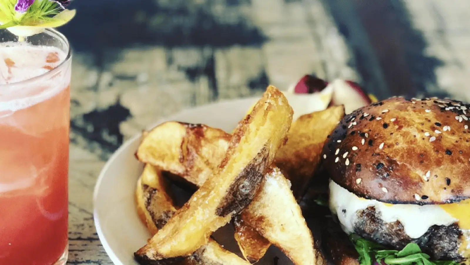 A plate with a gourmet burger featuring a sesame seed bun, melted cheese, and fresh greens, served with thick-cut potato wedges. Next to the plate is a reddish beverage garnished with a small flower and a citrus slice. The background is a weathered wooden table, perfect for the best dinner on the Monterey Peninsula.