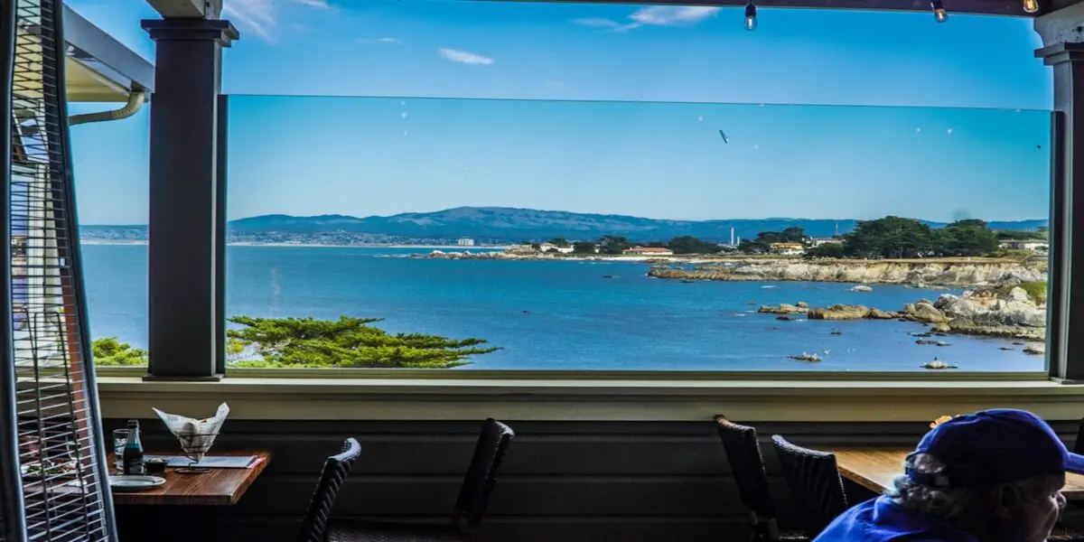 A scenic view of a coastal landscape from a covered patio with dining tables at one of the best restaurant views on the Monterey Peninsula. The ocean is calm, with rocky cliffs and trees along the shoreline. Clear blue sky with a few clouds and distant mountain range. Partial view of a person seated in the foreground.