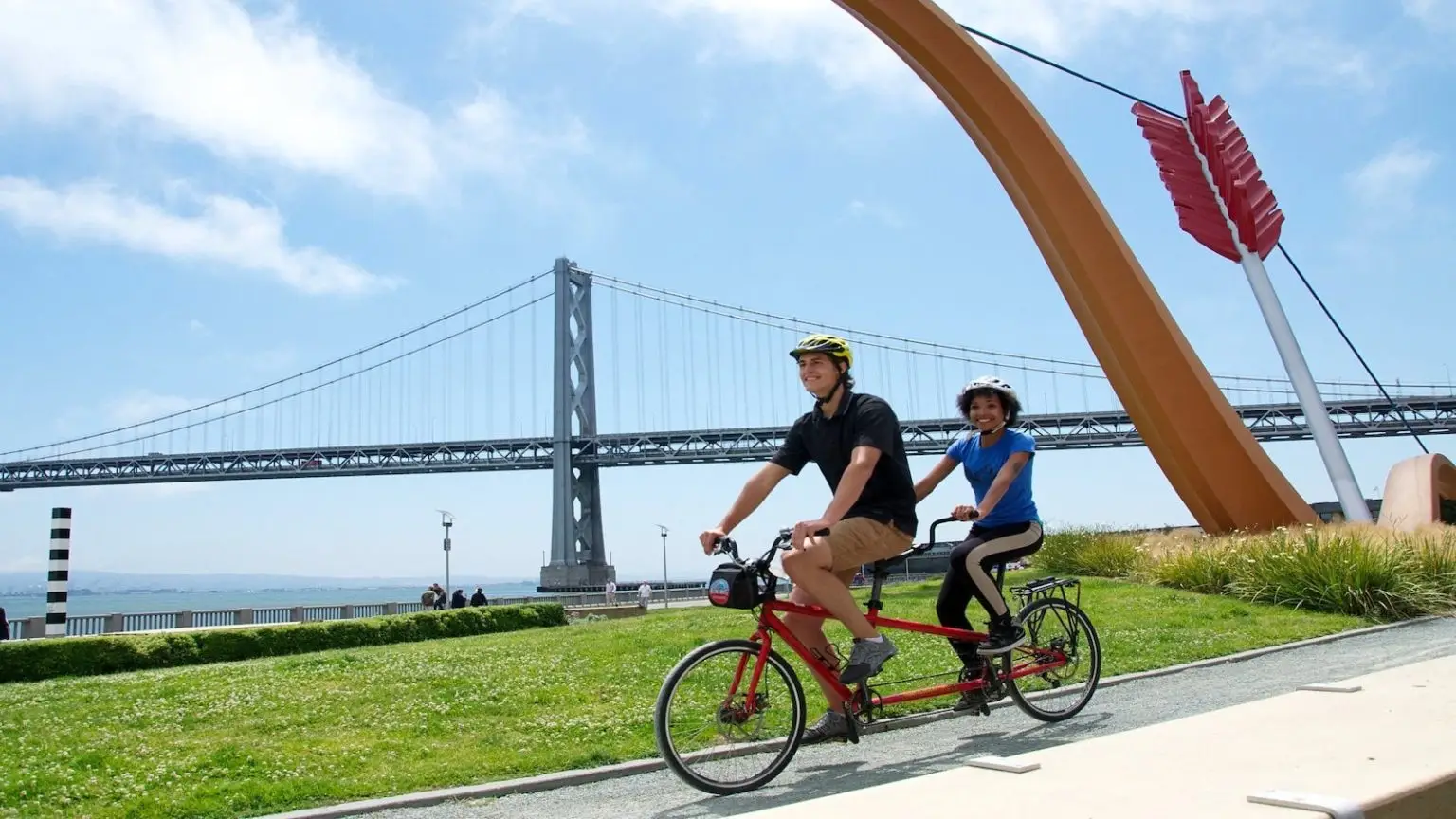 Two people ride a red tandem bicycle near a large arrow sculpture in a park. In the background, a suspension bridge spans across the blue sky with scattered clouds. The riders, perhaps discussing Top Food Tours in San Francisco, wear helmets and enjoy a sunny day. Green grass and a walking path surround them.